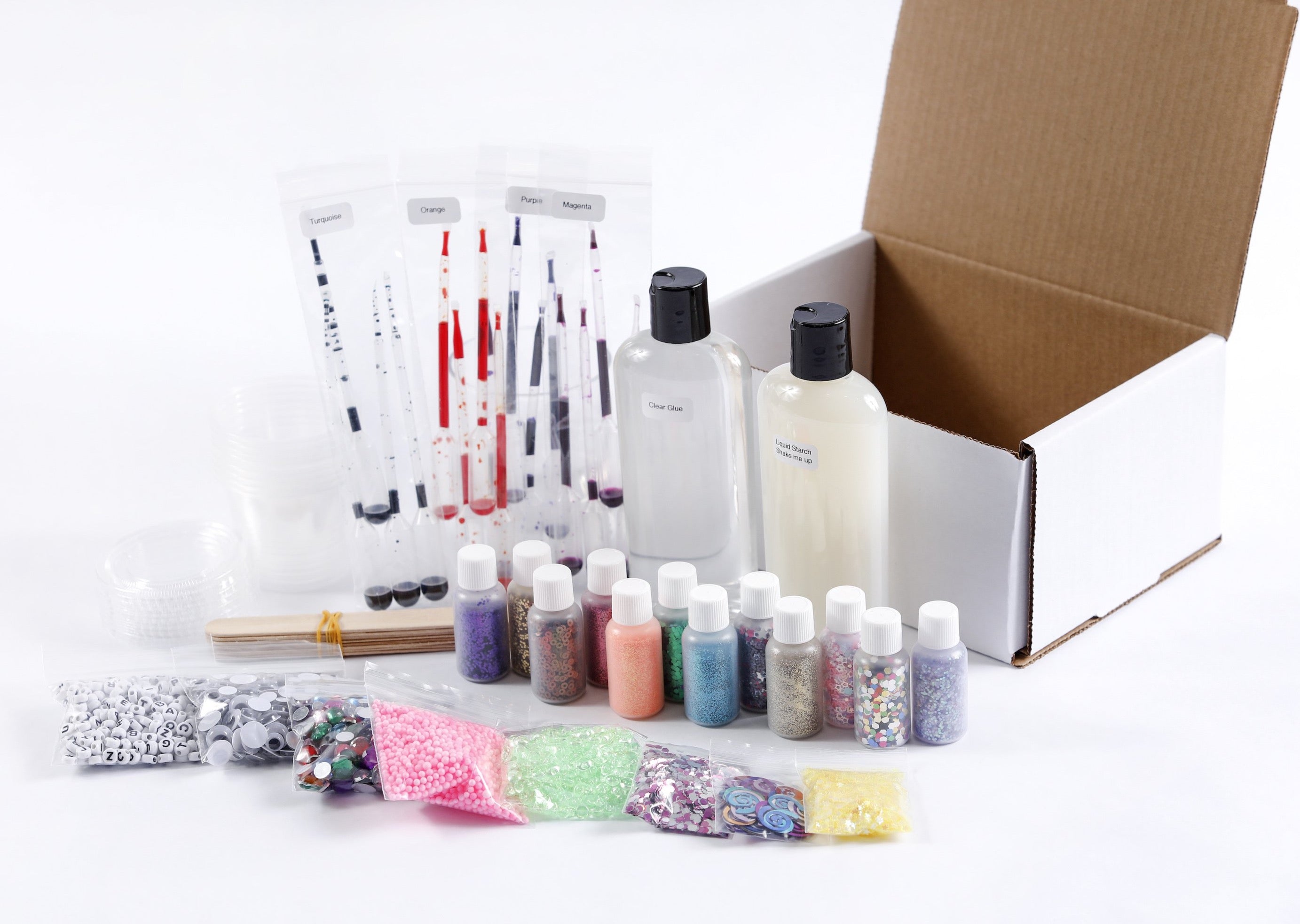 The slime making supplies on a table next to their empty box. Supplies include a bottles of glu, liquid starch, pipettes of colors, mixing containers, stir sticks. Plus extra mix-ins like glitter, beads, jewels, and googly eyes.