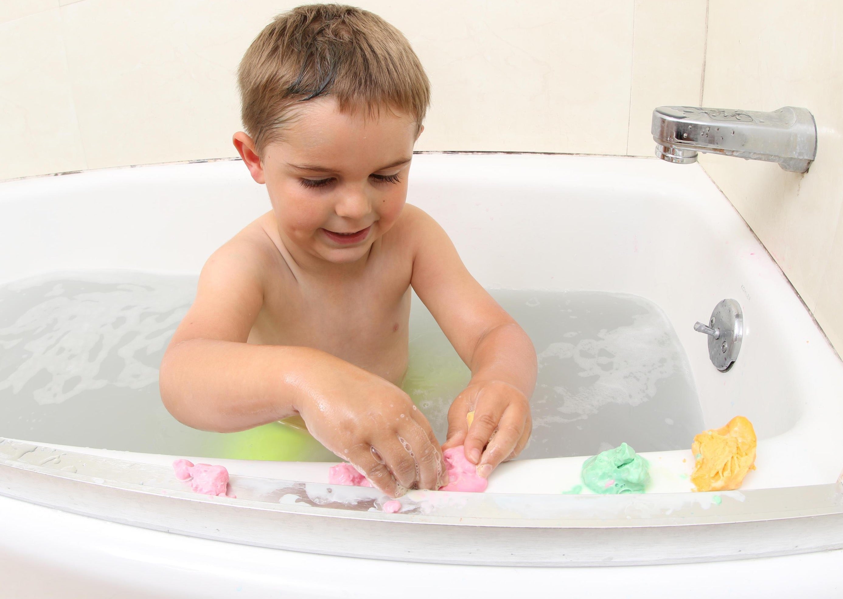 Child playing and molding playdough soap on the edge of a bathtub. Three colors of play dough soap including magenta, green, and yellow,