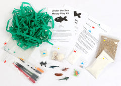 Materials and contents of the Under The Sea Messy Play Kit, including instructions, sand, corn starch, color tablets, toy fish, seashells, water beads, pipettes of watercolors, art paper, foil, and green ribbon seaweed.
