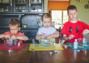 Three children standing next to a table making slime in three bowls on top of a colored mat.