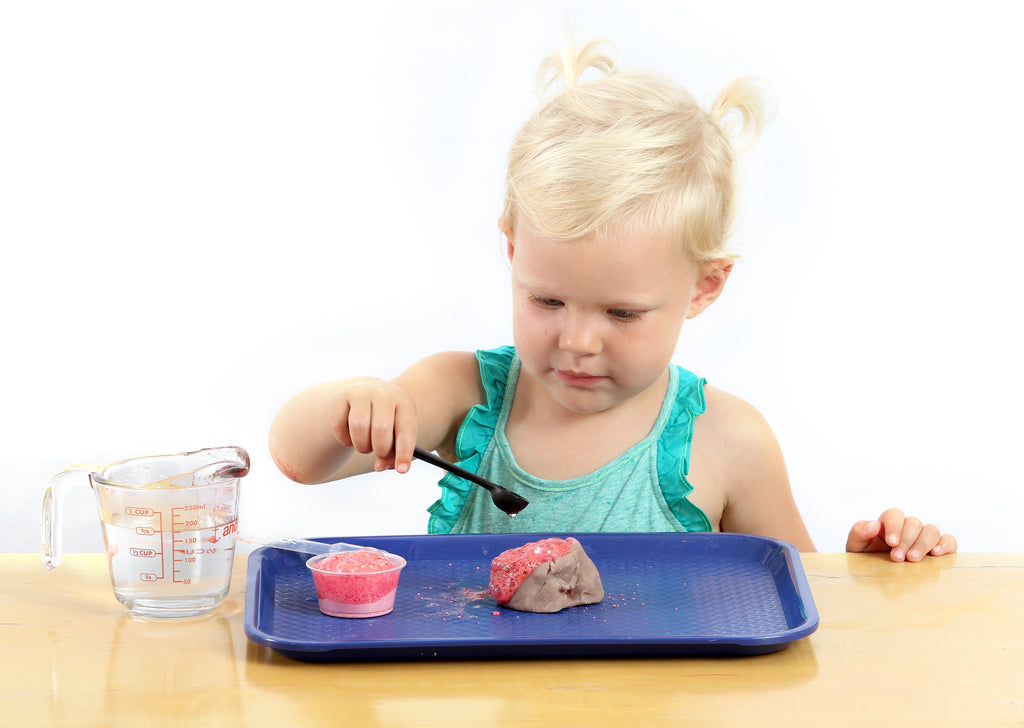 Girl concentrating on using a teaspoon to pour liquid over a clay volcano making it erupt with red lava.
