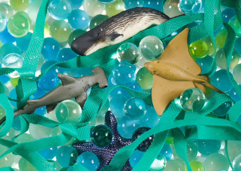 Close up of green and blue waterbeads in green ribbon seaweed with a toy shark, whale, and sting ray in it.