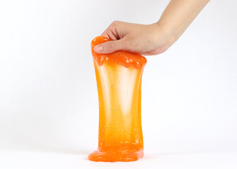 Hand holding Messy Play Kit's orange glitter slime stretching down towards the white tabletop.