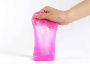 Hand holding Messy Play Kit's magenta pink glitter slime stretching down towards the white tabletop.