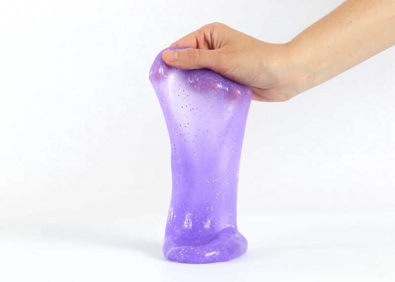 Hand holding Messy Play Kit's purple glitter slime stretching down towards the white tabletop.