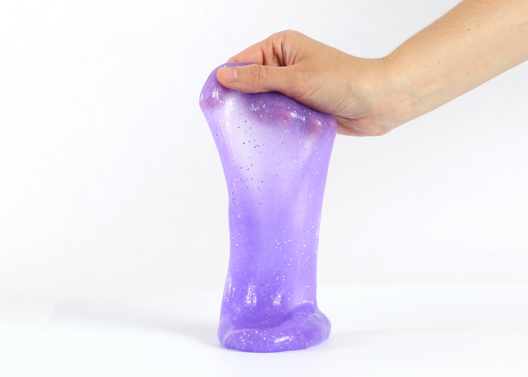 Hand holding Messy Play Kit's purple glitter slime stretching down towards the white tabletop.