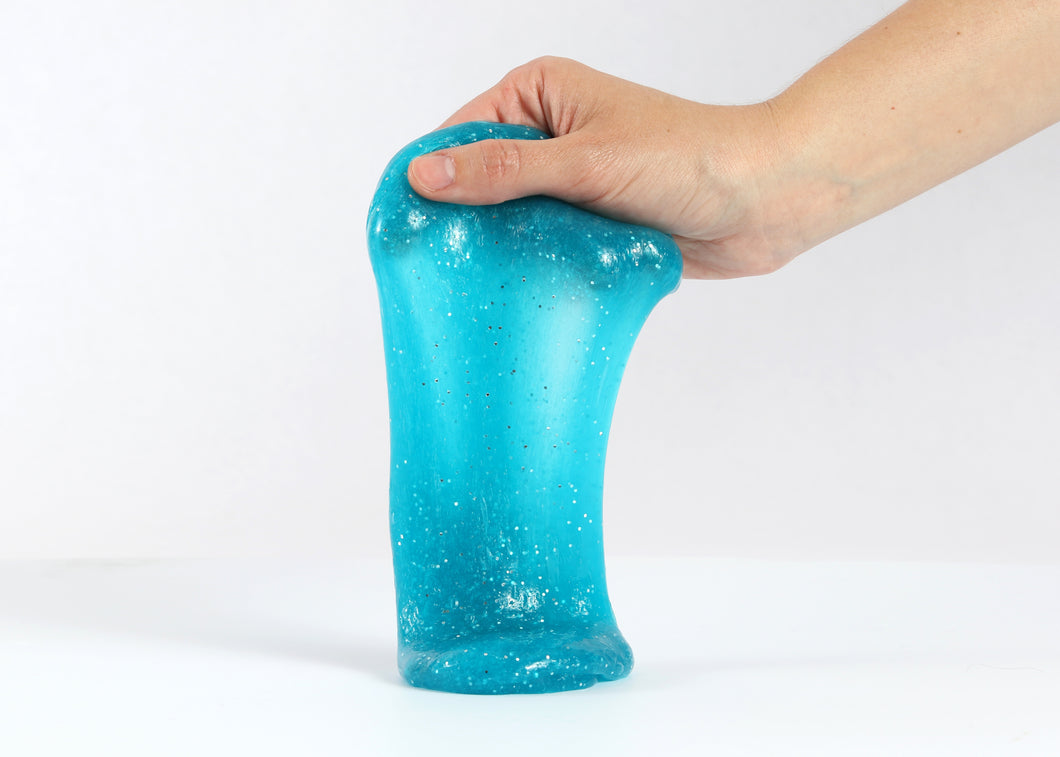 Hand holding Messy Play Kit's turquoise blue glitter slime stretching down towards the white tabletop.