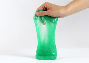Hand holding Messy Play Kit's green glitter slime stretching down towards the white tabletop.