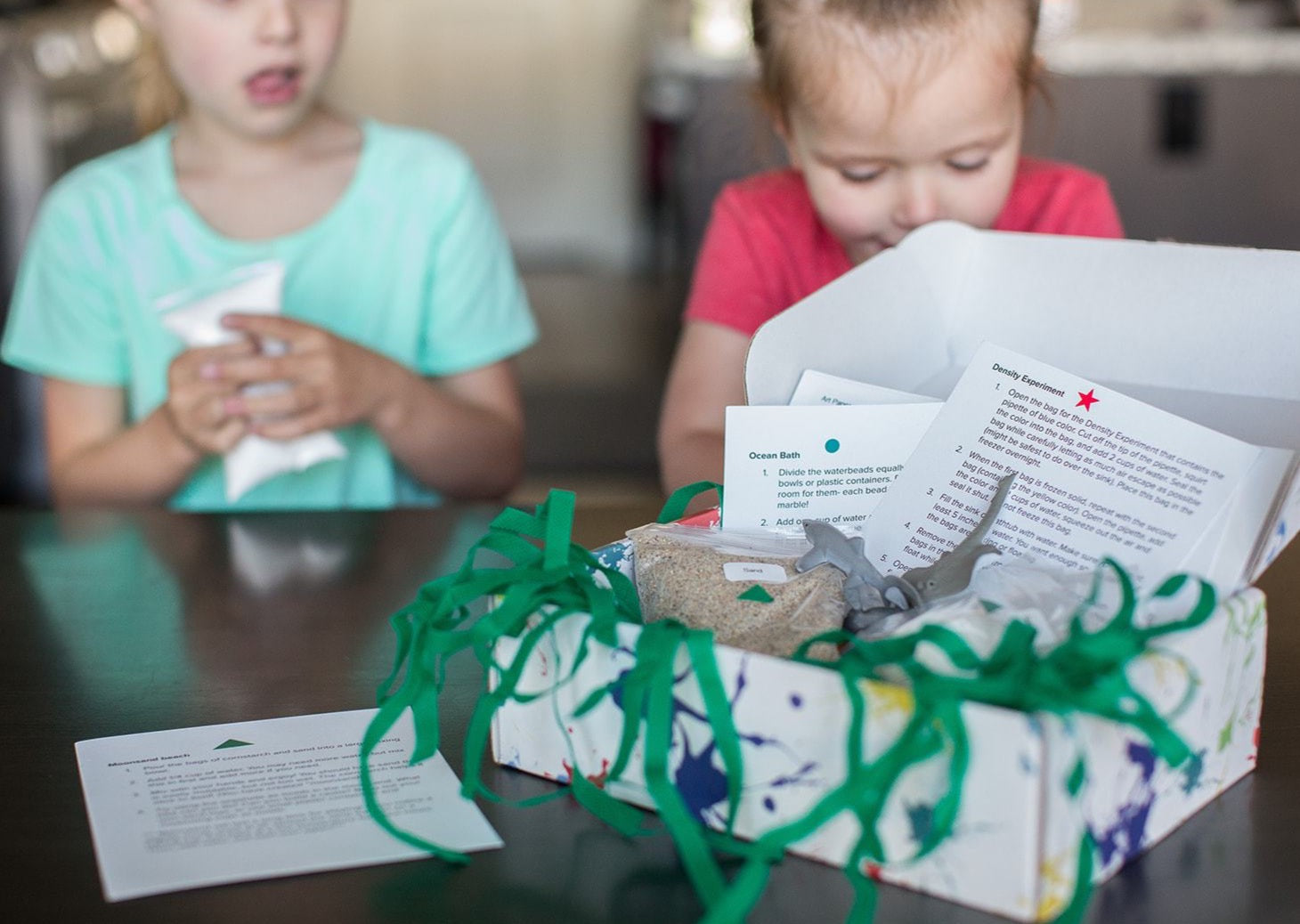 Two girls looking at the box of the Under The Sea Messy Play Kit with the instructions and seaweed coming out of it.