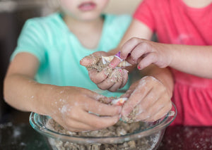 Children's hands digging seashells out of a glass bowl of sand on a table.