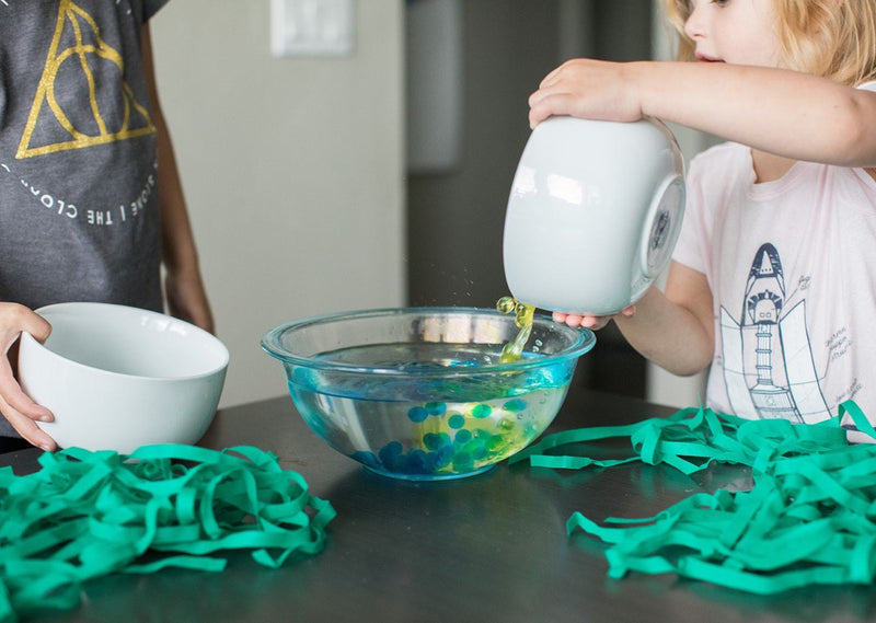 Child pouring waterbeads into a bowl of water with seaweed on the table.