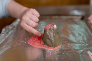 Child's hand pointing at a play dough volcano that has erupted red lava