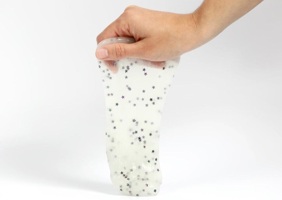 Hand holding Messy Play Kit's glow-in-the-dark slime in a bright room. Slime is white with glitter stars.