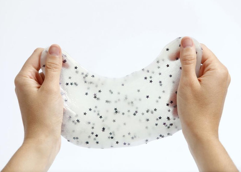 Two hands in a bright room pulling and stretching the glow in the dark slime. Slime is white with glitter stars.