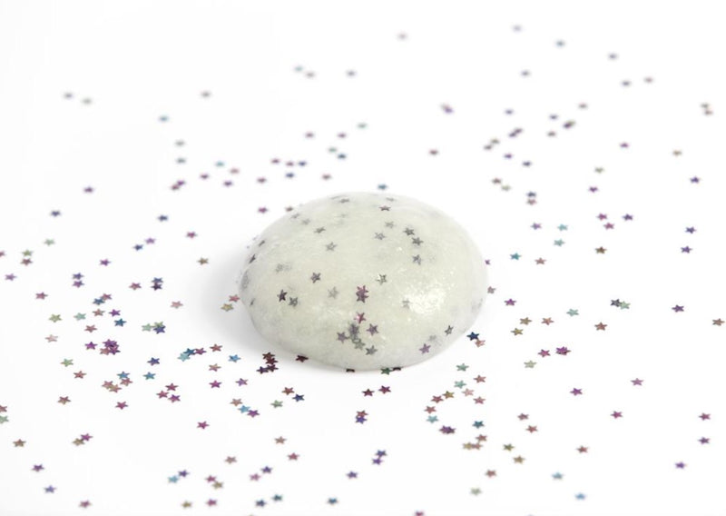 A ball of white Glow in the Dark slime with glitter stars laying on a table in a bright room surrounded by glitter.