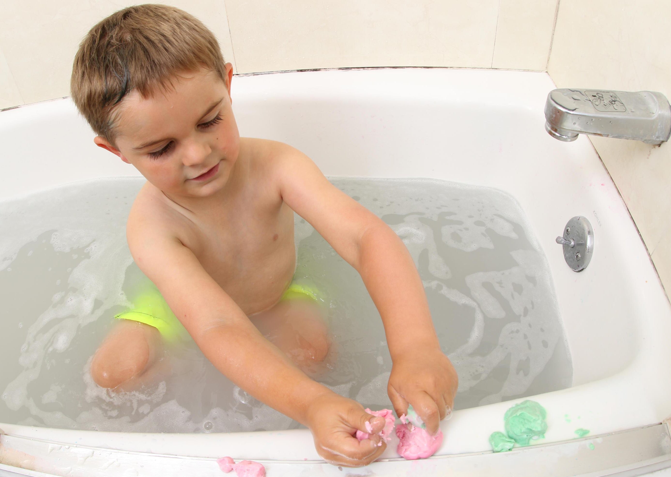 Child playing with and breaking apart playdough soap on the edge of a bathtub.